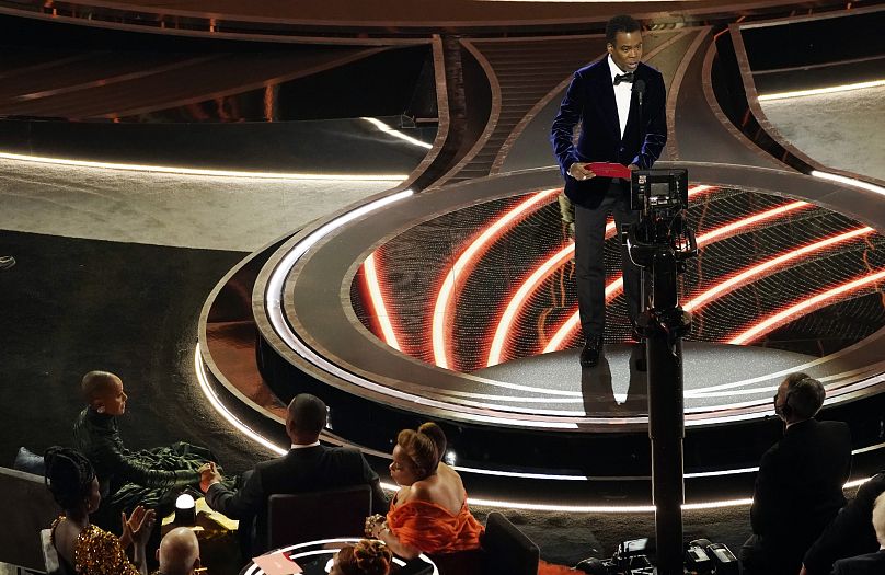 Aux Oscars 2022, le 27 mars, le présentateur Chris Rock monte sur scène sous les yeux de Jada Pinkett Smith et Will Smith, qui le confronteront plus tard, depuis leurs sièges.