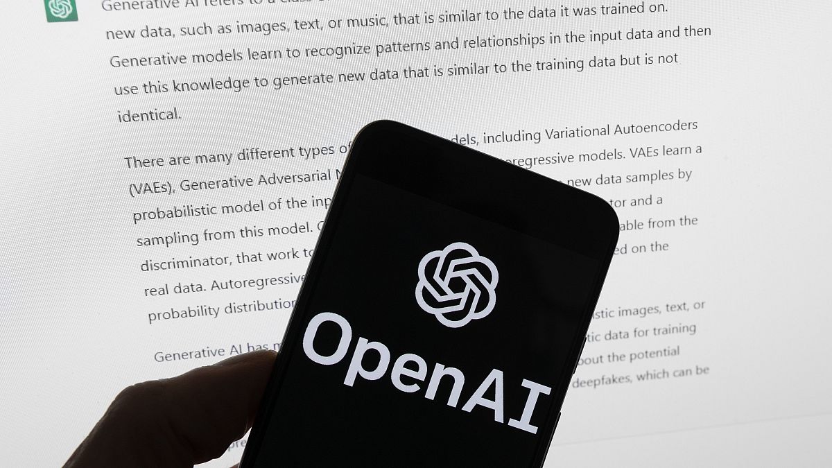 The OpenAI logo is seen on a mobile phone in front of a computer screen displaying output from ChatGPT, March 21, 2023, in Boston.