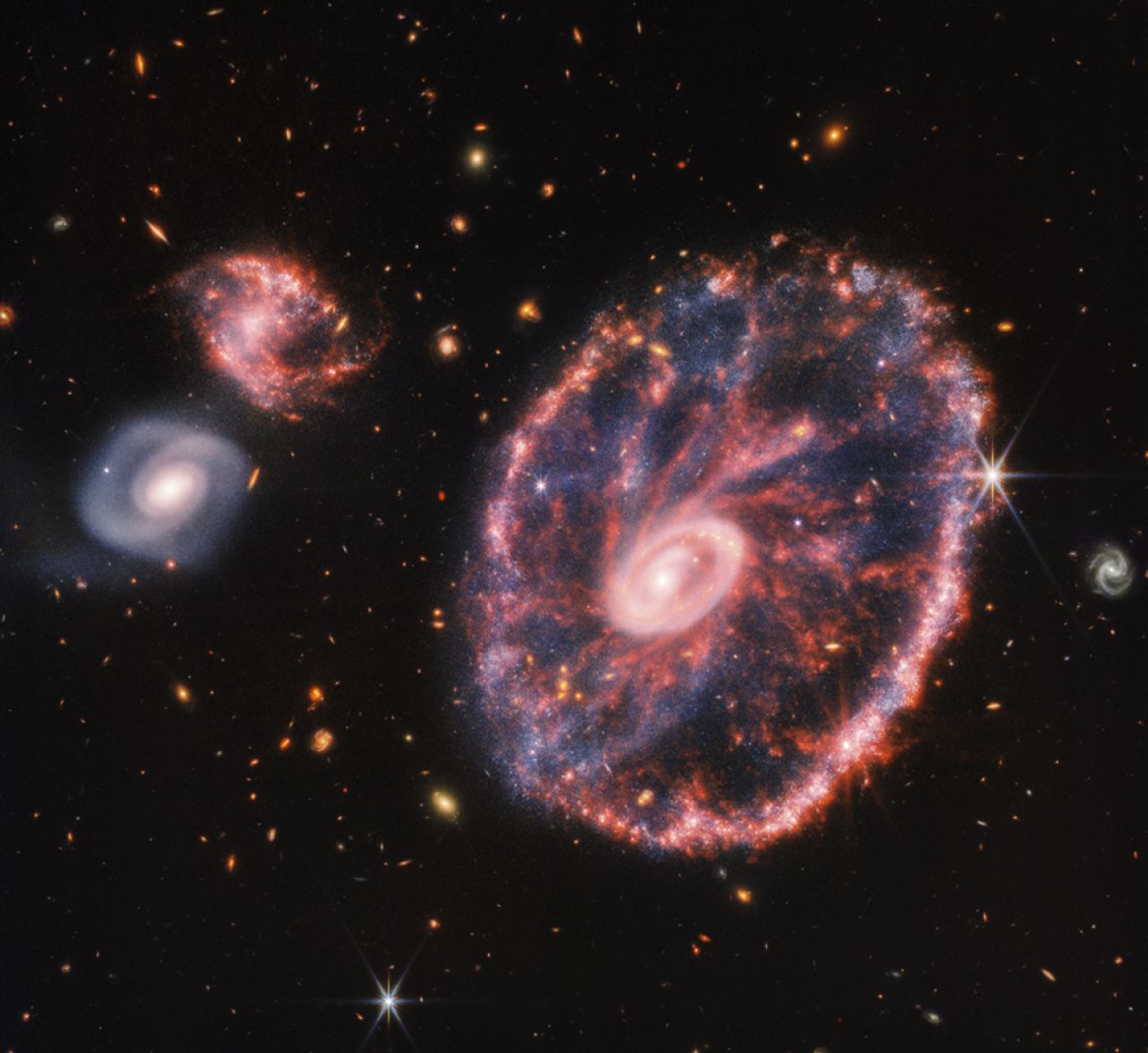 une grande galaxie rose tachetée ressemblant à une roue avec un petit ovale intérieur, avec du bleu poussiéreux entre les deux à droite, avec deux galaxies spirales plus petites à peu près de la même taille.