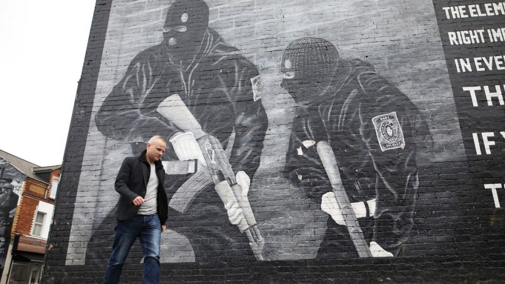 Loyalist activist Jamie Bryson walks past a Ulster Volunteer Force mural in east Belfast, Northern Ireland, Tuesday, Oct. 15, 2019.