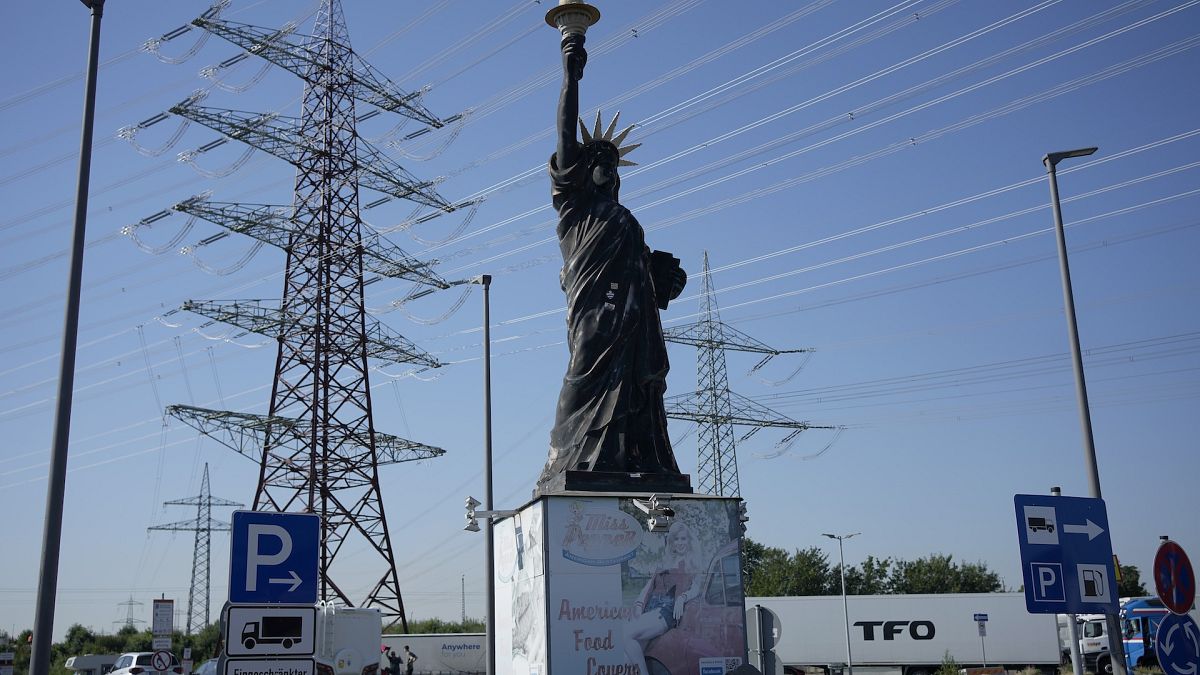 igh voltage pylons stand behind a replica of the statue of liberty, at a petrol station where trucks are parked near Cologne