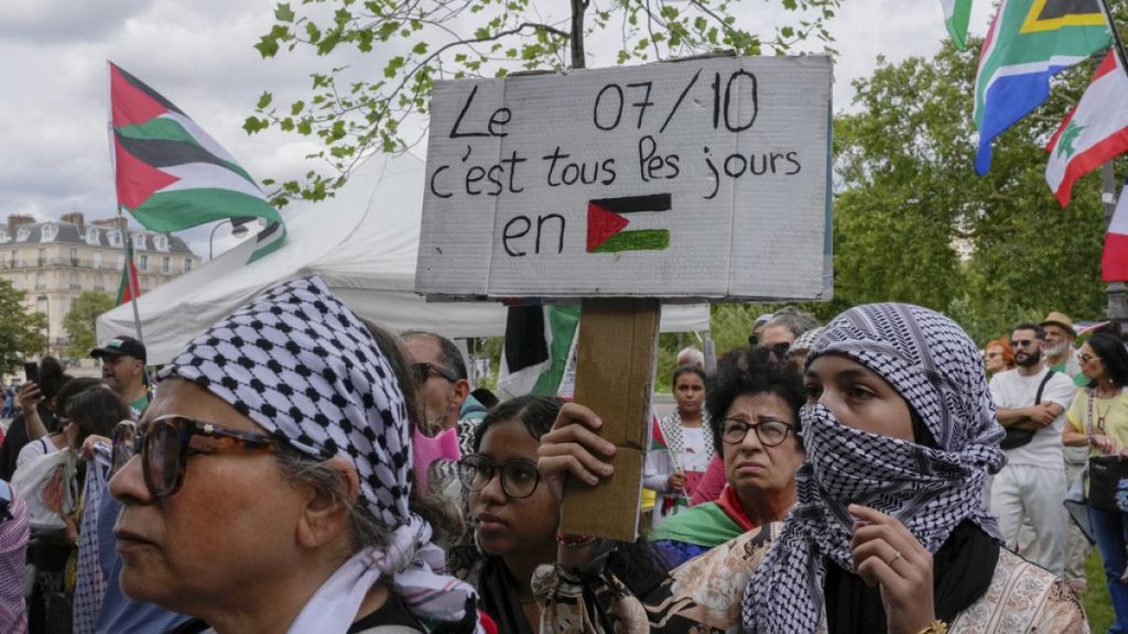 A protester wearing a keffiyeh holds a placard that reads,