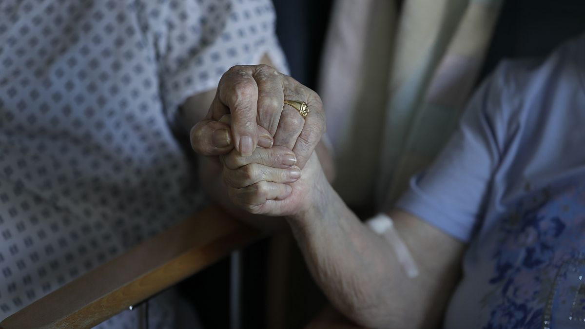 An elderly couple holds hands during a visit.